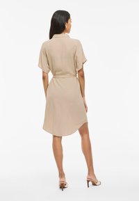 Beige skjortklänning med knytbar midja, korta ärmar och en rundad nederkant. Tillverkad av lätt tyg med en mjuk textur.