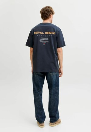 Moški, ki nosi temno modro majico z napisom "Royal Denim Division", modre kavbojke in bež čevlje, stoji obrnjeni stran na enostavni podlagi.