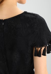Vestido negro con un patrón de paisley texturizado, mangas cortas con acentos de flecos negros y cierre de cremallera en la parte posterior.
