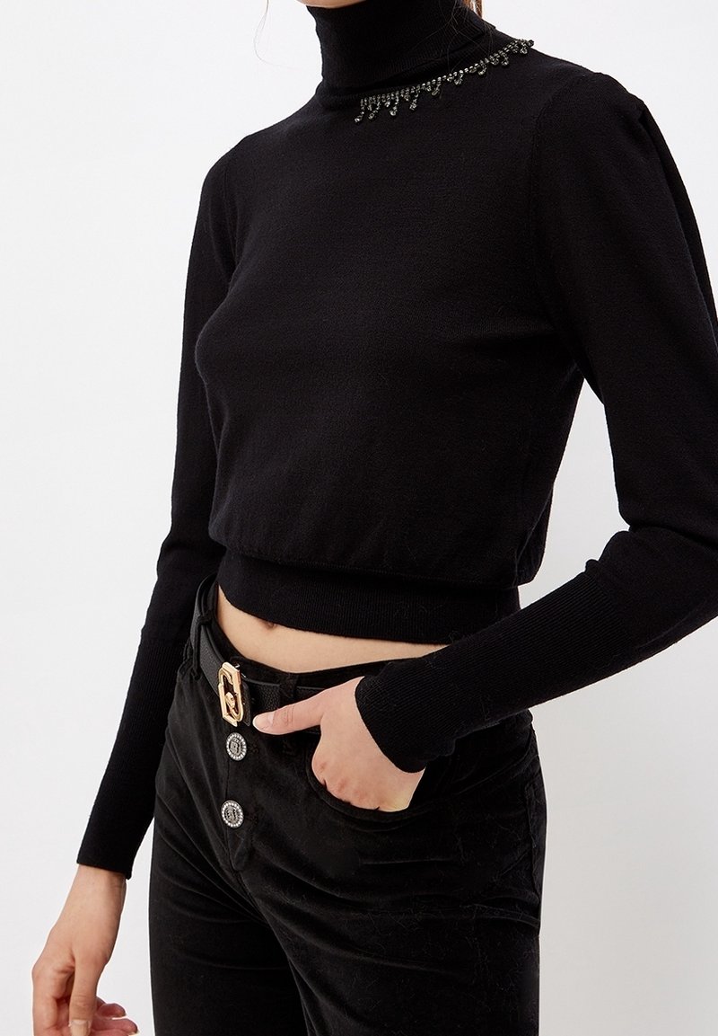 Femme portant un pull court noir à col roulé avec des détails en perles sur le col, un pantalon noir avec trois boutons décoratifs et une ceinture.