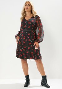 Robe noire avec motif floral rouge, manches longues transparentes, décolleté en V, clous décoratifs à la taille et ourlet à volants. Associée avec des bottines noires.