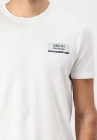 Baltos trumpomis rankovėmis marškinėliai, pagaminti iš medvilnės. Turi stačiakampio formos logotipo pleistrą šviesiai žaliai ir tamsiai mėlynos spalvos, su tekstu "DENIM TOM TAILOR."