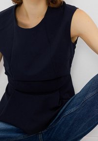 Person wearing a sleeveless dark navy top and blue jeans, sitting with legs crossed against a plain white background.
