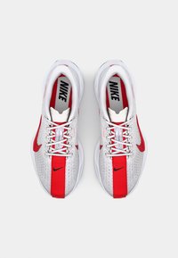 Baskets de sport blanches avec des accents rouges, tige en mesh, design à lacets, branding Nike en évidence et semelles texturées pour l'adhérence.