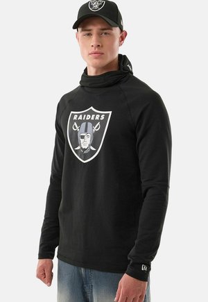 Młody mężczyzna noszący czapkę Raiders w kolorze czarnym i czarną koszulkę z długim rękawem z logo Raiders, w połączeniu z jasnoniebieskimi dżinsami, stojący na jednolitym tle.