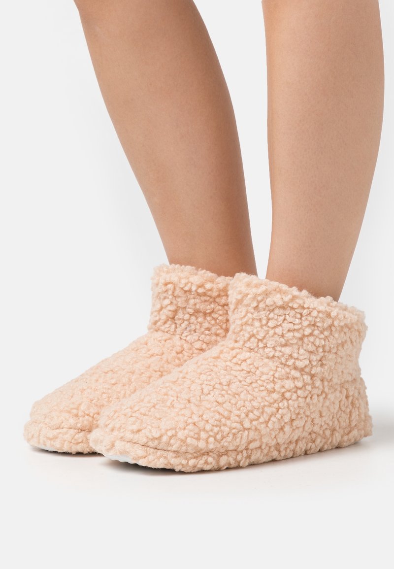 Pieds portant des chaussons en polaire beige clair, doux et texturés, avec un col montant au niveau de la cheville sur fond blanc.