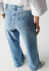 Jean large bleu clair avec taille haute, comprenant deux poches arrière et une étiquette de marque visible sur la ceinture. Texture en denim douce.