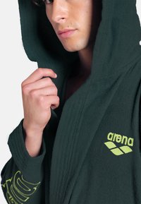 Accappatoio con cappuccio in spugna verde scuro, con una finitura testurizzata e un logo giallo con un design grafico sulla manica.