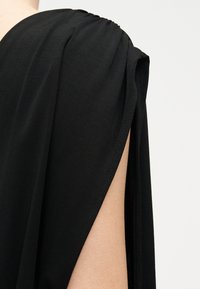 Κοντινό πλάνο ενός ώμου που φοράει μαύρο τοπ χωρίς μανίκια με φαρδιά μασχάλη και λεπτομέρεια υφάσματος που πέφτει, σε απλό φόντο.