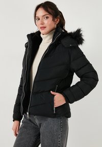 Crna puffer jakna s otpojenim lažnim krznom na kapuljači, zatvorenim prednjim dijelom s patentnim zatvaračem i bočnim džepovima, koja ima kviltanu teksturu i uski kroj.
