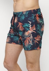 Costume de baie dintr-un material bleu închis, cu un model vibrant de frunze tropicale în verde și portocaliu, cu un elastic de ajustare la talie și detalii de branding pe lateral.