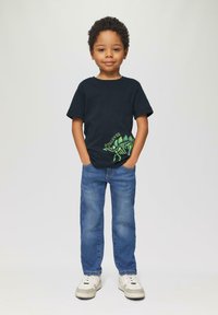 Marineblaues T-Shirt mit einem grünen Dinosaurier-Design und Text, kombiniert mit hellblauen Jeans und weißen Sneakers mit grauen Akzenten.