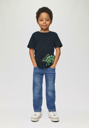 Marineblauw T-shirt met groene dino-afbeelding en tekst, gecombineerd met lichtblauwe denim jeans en witte sneakers met grijze accenten.