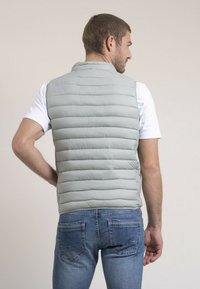 Gilet rembourré en tissu gris clair, avec un matelassage horizontal et un col haut. Porté sur un t-shirt blanc, associé à un jean bleu.