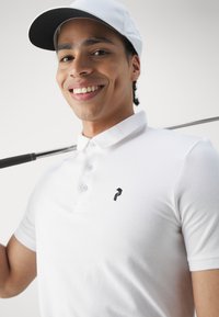 Vit pikétröja med tre knappar och en svart logotyp. Slät bomullstextur, korta ärmar, bärs med en vit keps och en golfklubba.
