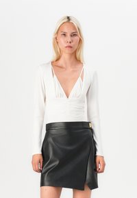 Long-sleeved white top with deep V-neck and gathered detail, paired with a black faux leather mini skirt featuring a side slit and gold hardware.