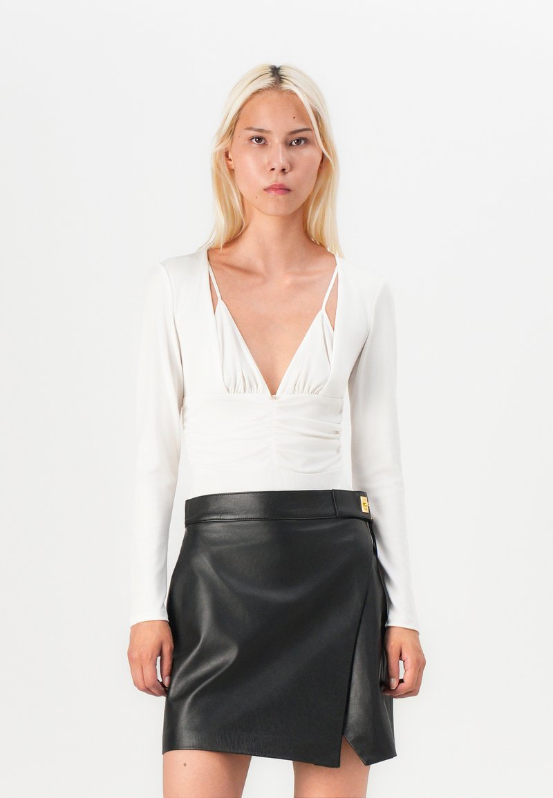 Long-sleeved white top with deep V-neck and gathered detail, paired with a black faux leather mini skirt featuring a side slit and gold hardware.