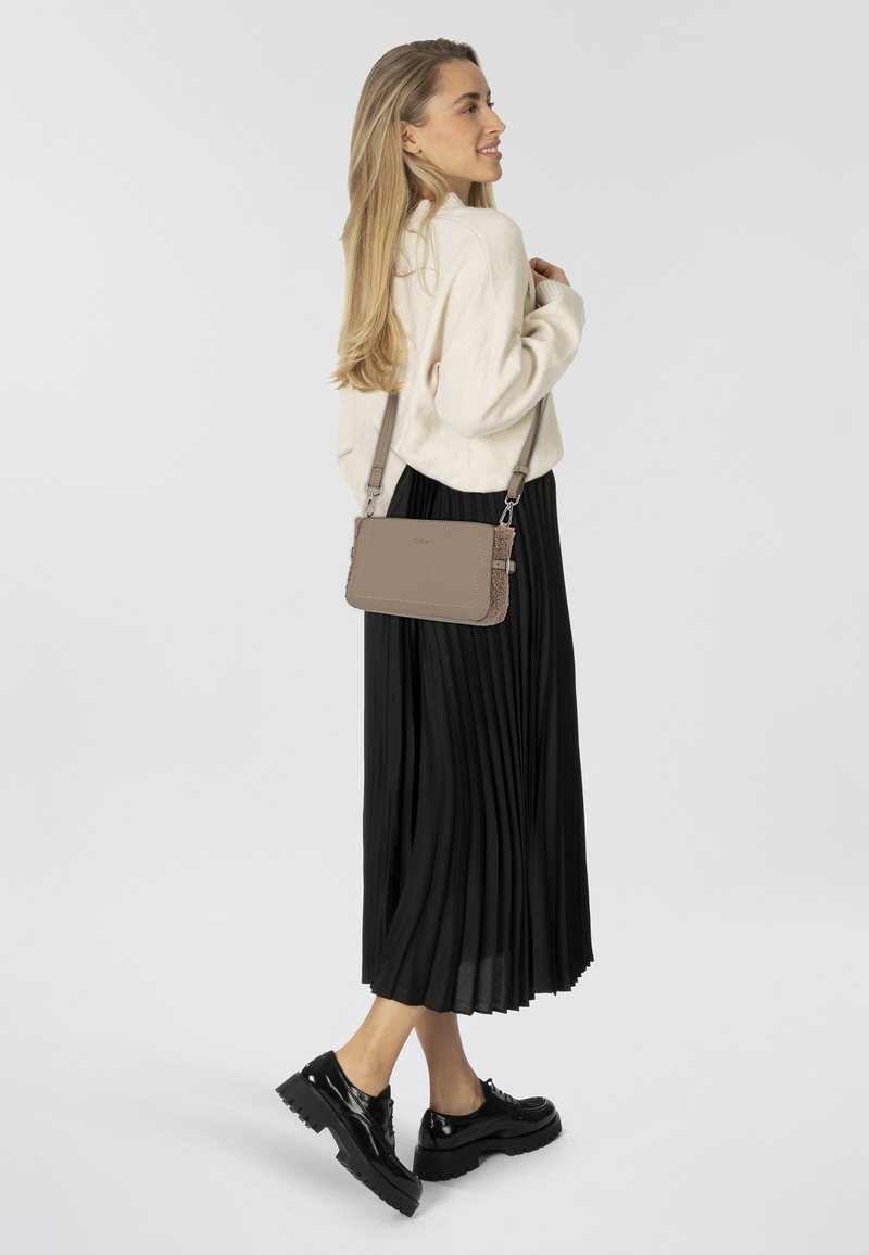 Borsa a tracolla in pelle beige con cuciture dettagliate, una texture liscia e hardware in argento, indossata sopra una gonna plissettata nera e scarpe.