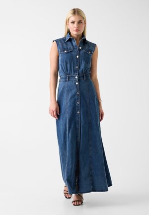 Femme blonde portant une robe maxi en denim sans manches avec boutons sur le devant et des sandales noires à brides, debout sur fond blanc.