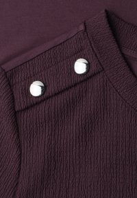 Gros plan d’un tissu texturé violet foncé avec des boutons-pression argentés sur une couture d’épaule pliée.