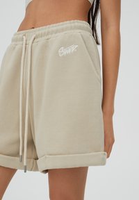 Shorts en coton beige avec des revers, taille ajustable à cordon, poches latérales et un petit logo blanc brodé à l'avant.