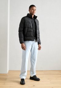 Svart dunjakke med høy krage, to frontlommer og kviltet tekstur, paret med lyseblå rette jeans og svarte sko.