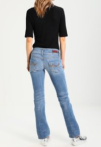 Jeans acampanados de mezclilla azul claro con bolsillos traseros que presentan detalles de costura. Combinados con una camiseta negra de manga corta y zapatos plateados.