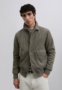 Chaqueta de pana en verde olivo, con cuello, dos bolsillos frontales, cierre de botones y puños y cintura acanalados. Textura suave.