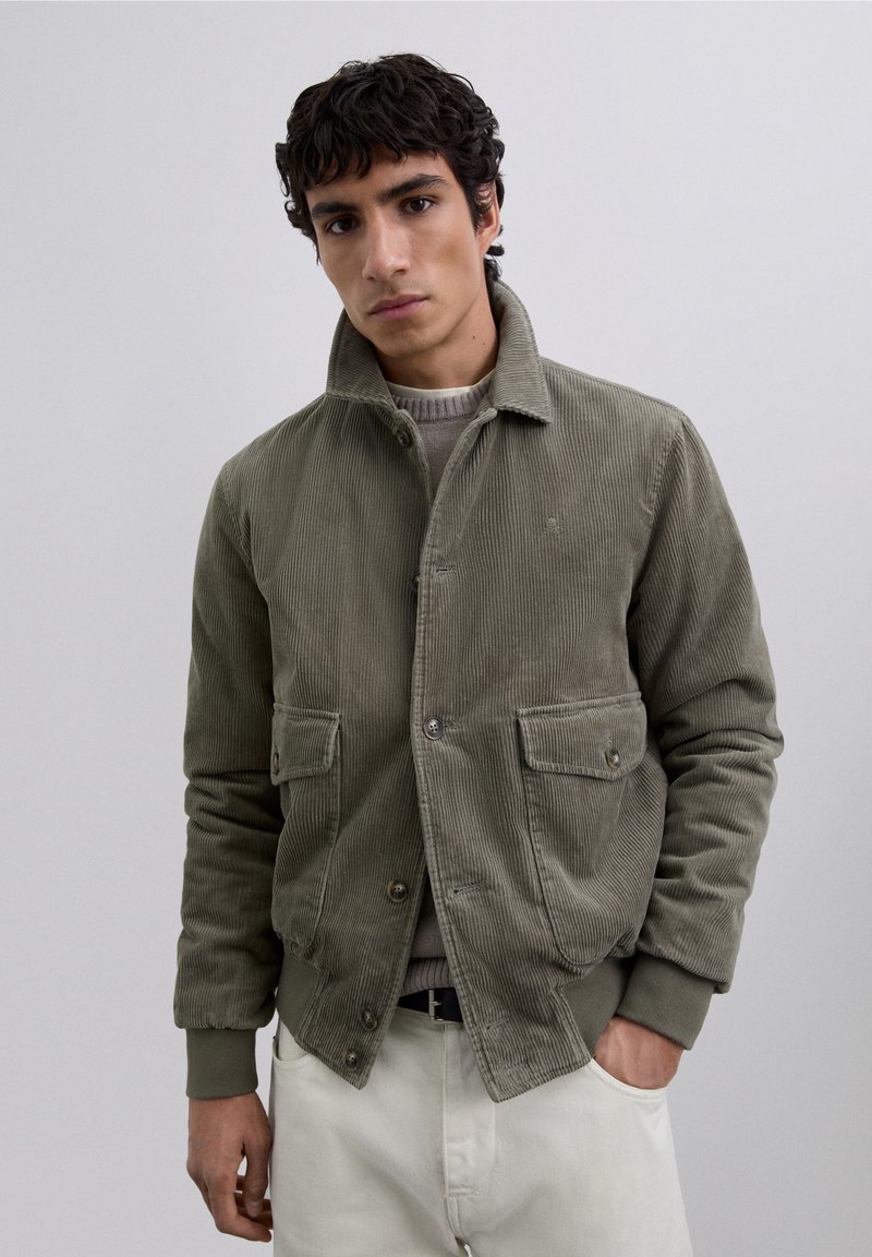 Chaqueta de pana en verde olivo, con cuello, dos bolsillos frontales, cierre de botones y puños y cintura acanalados. Textura suave.