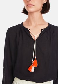 Schwarze Langarmbluse mit gerafftem Ausschnitt, ausgestattet mit einer weißen Schnur und orange-weißen Quasten als Akzenten. Leichter, luftiger Stoff.