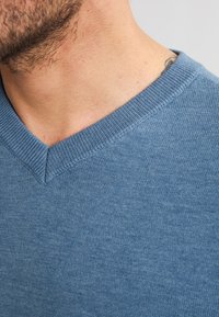 Pulover cu decolteu în V, realizat dintr-un material moale, texturat de culoare albastră. Prezintă detalii ribbate în jurul gulerului și o formă ajustată.