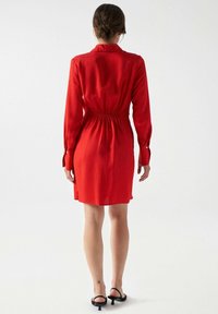 Robe rouge à manches longues avec un corsage ajusté, taille élastique, tissu lisse et ourlet au genou. Des sandales noires complètent le look.