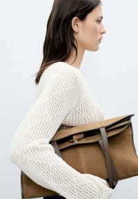 Geantă din suede maron cu accente din piele, design rectangular și închidere cu clapetă superioară. Pur tată cu un pulover tricotat crem.