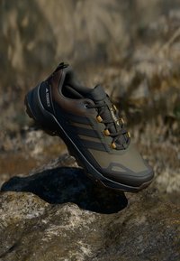 Terrex Wanderschuh mit einem obermaterial in dunklem Grün und Braun, strukturiertem Material, gelben Schnürsenkeln und einer robusten Sohle, die für den Einsatz im Freien konzipiert ist.