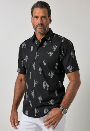 Hombre con cabello corto y oscuro y barba, vestido con una camisa negra de manga corta con estampados blancos de cactus y pantalones blancos sobre un fondo liso.