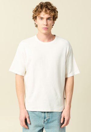 Joven con cabello rizado, viste una camiseta blanca de manga corta y unos jeans azul claro, frente a un fondo neutro.