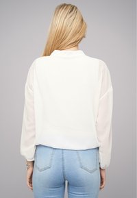 Hvid bluse med gennemsigtige ærmer, rund halsudskæring, og en rygkant, der let buer. Kombinationen er med tætsiddende lyseblå jeans.