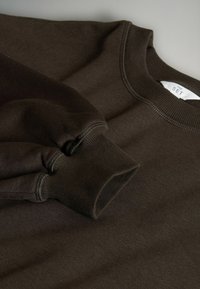 Hnědá mikina s kulatým výstřihem a ribovanými manžetami a límcem. Měkká textilie a čisté švy, s etiketou na vnitřním límci.