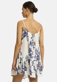 Das Modell steht mit dem Rücken zur Kamera und trägt ein ärmelloses Blumenkleid mit einem Rüschensaum, das auf einem hellen Hintergrund blaue Muster aufweist.