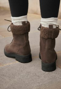 Botas de tobillo de ante marrón con un puño de borreguillo marrón y esponjoso, cordones en la parte delantera y una suela de goma negra texturizada. Medias de lana son visibles sobre el puño.