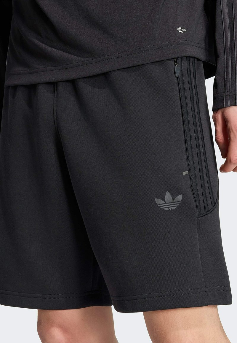 Pantaloncini sportivi neri con logo Adidas e tasca laterale con zip, indossati con top nero coordinato.