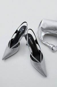 Zilveren slingback hakken met spitse neuzen, bedekt met een textuur van gaasstof. Zwarte binnenvoering, lage hak en een zilveren handtas ernaast.