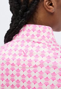 Prim-plan cu părul împletit al unei persoane și jumătatea superioară a spatelui, purtând o cămașă roz și albă cu modele în formă de evantai.