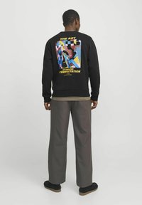 Sudadera negra con un diseño gráfico colorido en la parte posterior, que presenta formas abstractas y texto. Combinada con pantalones grises holgados y zapatos negros.