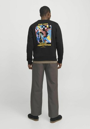 Zwarte sweatshirt met kleurrijk grafisch ontwerp op de achterkant, met abstracte vormen en tekst. Gepaard met losse, grijze broeken en zwarte schoenen.