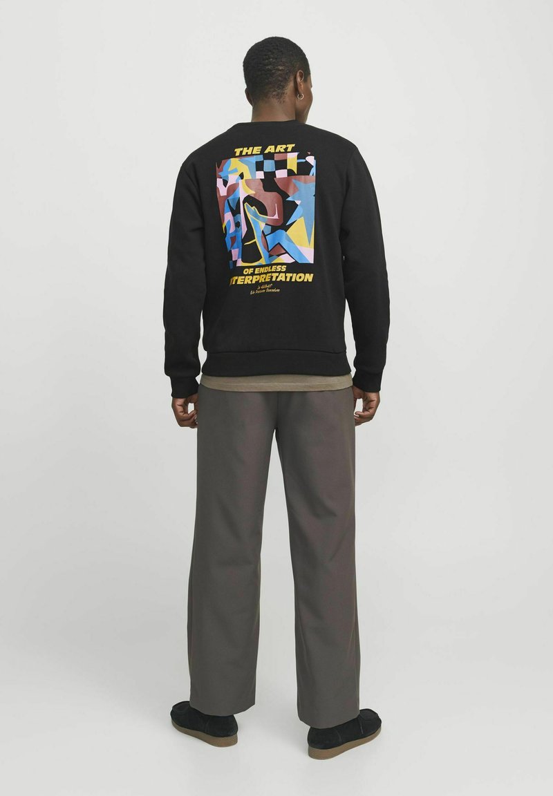 Sudadera negra con un diseño gráfico colorido en la parte posterior, que presenta formas abstractas y texto. Combinada con pantalones grises holgados y zapatos negros.