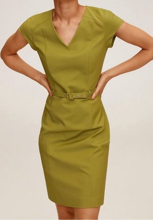 Frau trägt ein eng anliegendes olivgrünes Kleid mit kurzen Ärmeln, V-Ausschnitt und passendem Gürtel, steht mit den Händen in der Hüfte vor einem schlichten Hintergrund.