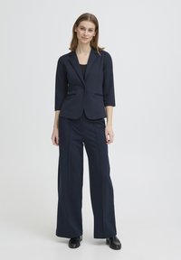 Blazer bleu marine à manches trois-quarts, fermeture à un seul bouton, et poches latérales, associé à un pantalon large assorti et des chaussures noires.