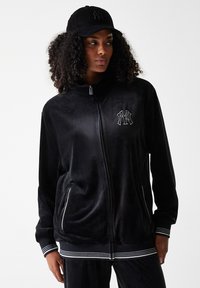 Veste en velours noir avec fermeture éclair et logo blanc, accompagnée de poignets rayés. Dispose de deux poches latérales et d'un col montant. Assortie d'une casquette noire.