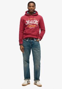 Rode hoodie met witte tekst en logo, blauwe geruite shirt eronder, blauwe spijkerbroek en witte sneakers met grijze details.